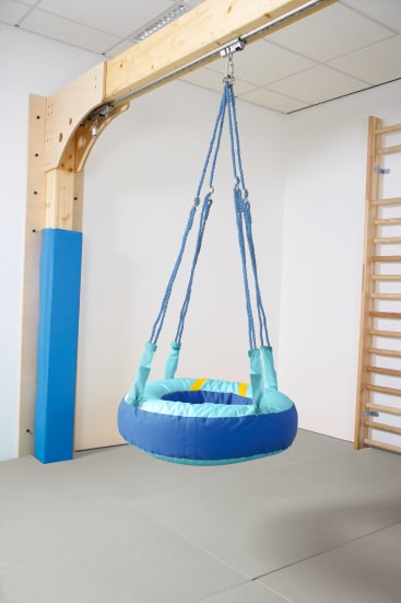 Eine gepolsterte Rundschaukel mit blauen Seilen hängt an einem Holzgestell in einem Raum mit grauem Teppichboden, einer Wandleiter und weißen Wänden.