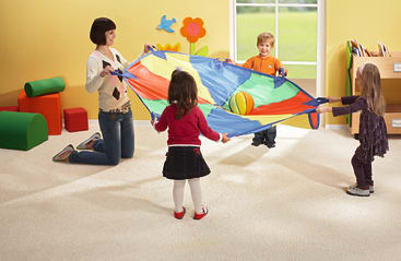 Ein Erwachsener und drei kleine Kinder spielen mit einem bunten Fallschirm und einem Ball in einem hellen, mit Teppich ausgelegten Klassenzimmer mit weichen Spielmöbeln, Bücherregalen und Wanddekorationen.