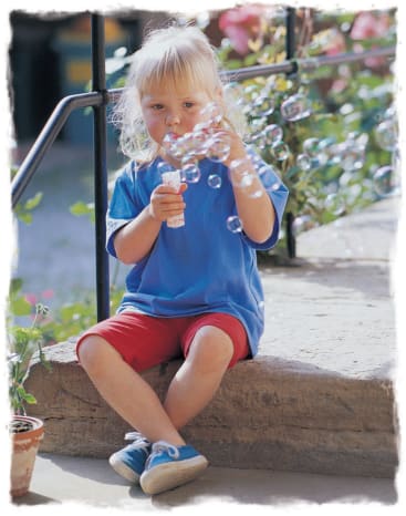 Ein kleines Kind mit blondem Haar, das ein blaues Hemd und rote Shorts trägt, sitzt auf einer Steinstufe im Freien und pustet Seifenblasen, während im Hintergrund grüne Pflanzen und Blumen zu sehen sind.