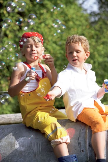 Zwei Kinder sitzen im Freien und pusten Seifenblasen. Das Kind auf der linken Seite trägt ein rotes Kopftuch und einen gelben Overall, während das Kind auf der rechten Seite ein weißes Hemd und orangefarbene Shorts ohne Umschlag trägt. Im Hintergrund sind grüne Bäume zu sehen.
