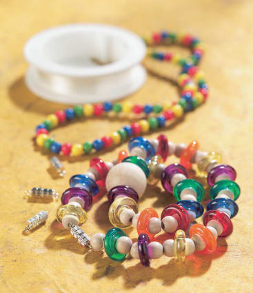 Bunter Perlenschmuck, darunter ein Armband und eine Halskette mit runden Perlen in verschiedenen Farben, ist auf einer gelben Fläche angeordnet. Eine Spule mit durchsichtiger Schnur und mehrere silberne Verschlüsse sind ebenfalls zu sehen.