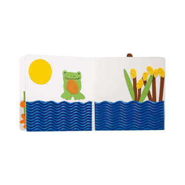 Eine bunte Filzillustration in einem Kinderbuch zeigt einen Frosch am Flussufer unter einer gelben Sonne, mit blauem, welligem Wasser und hohen grünen und braunen Rohrkolben auf der rechten Seite.