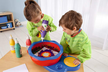 Zwei Kinder in hellgrünen Kitteln benutzen an einem Tisch eine Spinning-Art-Maschine. Ein Kind drückt Farbe aus einer Flasche auf ein sich drehendes Papier; in der Nähe stehen mehrere Flaschen mit Farbe und ein Blatt Papier.