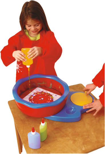Ein Kind in einem roten Kittel gießt gelbe Farbe in eine rote und blaue Kunstschleuder, während ein anderes Kind die Schleuder bedient. Flaschen mit Farbe stehen auf dem Holztisch.