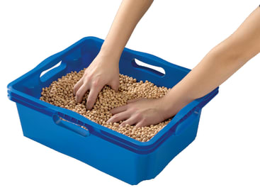 Zwei Hände sind in einen blauen Plastikeimer getaucht, der mit runden beigen Pellets gefüllt ist, wobei der Hintergrund entfernt wurde.