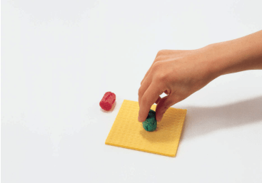 Eine Hand legt ein kleines grünes Objekt auf eine quadratische, gelbe, strukturierte Fläche, während ein kleines rotes Objekt in der Nähe auf einem weißen Hintergrund ruht.
