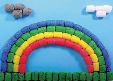 Bunte Bauklötze in Form eines Regenbogens mit fünf Bögen und einer grünen Basis vor blauem Hintergrund. Zwei Wolkenformen, eine graue und eine weiße, befinden sich am Himmel über dem Regenbogen.