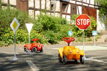 Zwei kleine Plastikspielzeugautos sind auf einer gepflasterten Fläche im Freien geparkt, umgeben von Verkehrsschildern, darunter ein Stoppschild und ein Vorfahrtsschild, mit Grünpflanzen und einem Gebäude im Hintergrund.