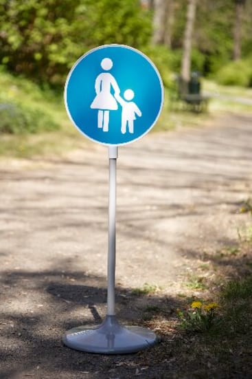 Ein blaues, rundes Schild mit weißen Symbolen eines Erwachsenen, der die Hand eines Kindes hält, ist auf einem weißen Ständer auf einem Kiesweg in einem Park im Freien mit Grün im Hintergrund angebracht.