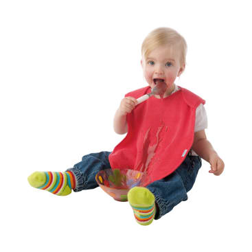 Ein Kleinkind, das ein rotes Lätzchen, eine blaue Jeans und bunt gestreifte Socken trägt, sitzt auf dem Boden und hält einen Löffel und eine Schüssel. Das Lätzchen ist mit Essen bekleckert, was auf eine unordentliche Essenssituation hindeutet.