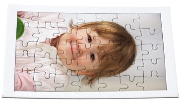 Ein fertiges Puzzle mit dem Foto eines kleinen Kindes mit hellbraunem Haar und einem rosa Hemd, das auf einer weißen Fläche liegt und in die Kamera schaut.