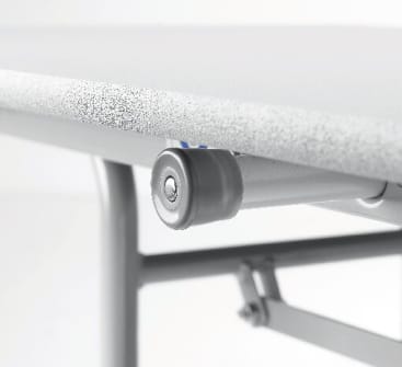 Nahaufnahme einer grauen Laufrolle, die unter einer strukturierten, hellen Tischplatte angebracht ist, wobei ein Teil des Metallrahmens und des Scharniermechanismus sichtbar ist.