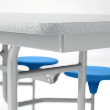 Nahaufnahme der Ecke eines grau gesprenkelten Tisches mit aufgesetzten runden Sitzen in Blau, Gelb, Rot und Grün, die von einem Metallrahmen getragen werden. Der Hintergrund ist einfarbig und weiß.