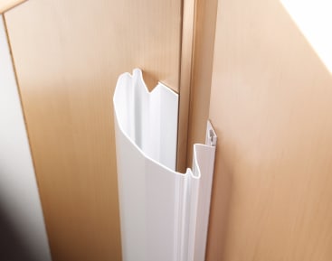 Ein weißer Kunststoff-Eckenschutz mit einer gezackten, gebrochenen Kante wird an der Ecke von Schränken aus hellem Holz angebracht.