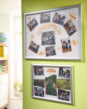 Zwei gerahmte Fotocollagen hängen an einer grünen Wand. Der obere Rahmen zeigt Klassen- und Gruppenfotos, die mit handschriftlichen Notizen beschriftet sind, während der untere Rahmen Gruppenfotos im Freien von Schülern auf einer Klassenfahrt zeigt.