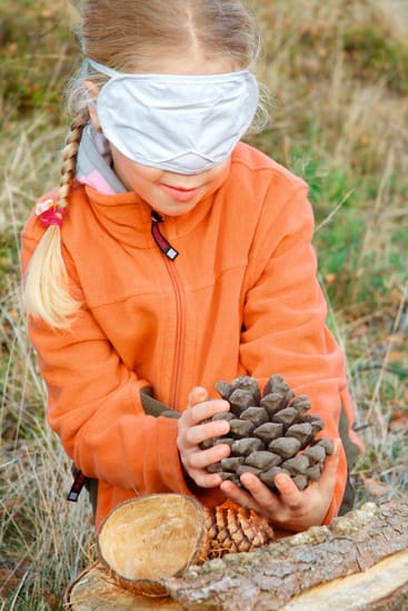 Ein junges Mädchen, das eine Augenbinde und eine orangefarbene Jacke trägt, hält mit beiden Händen einen großen Tannenzapfen im Freien, in der Nähe von geschnittenem Holz und Gras.