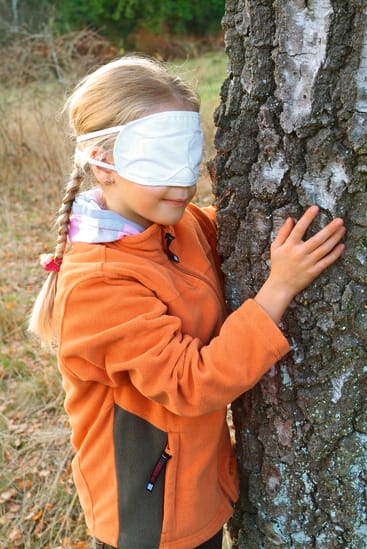 Ein junges Mädchen mit geflochtenem Haar, das eine orangefarbene Jacke und eine weiße Augenbinde trägt, steht im Freien und berührt den Stamm eines Baumes. Der Hintergrund ist grasbewachsen mit spärlicher Vegetation.