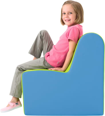 Ein kleines Kind sitzt seitlich auf einem blauen und grünen Schaumstoffstuhl, trägt ein rosa Hemd, eine graue Hose und rosa Slipper, schaut in die Kamera und lächelt.