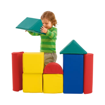 Ein kleines Kind in einem grün gestreiften Hemd legt einen großen grünen Schaumstoffblock auf eine bunte Struktur aus roten, gelben und blauen Schaumstoffblöcken vor einem weißen Hintergrund.