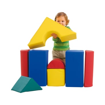 Ein kleines Kind in einem grünen Hemd steht hinter großen Schaumstoffbausteinen in den Farben Rot, Blau, Gelb und Grün und hält einen gelben dreieckigen Block über dem Bauwerk. Der Hintergrund ist einfarbig weiß.