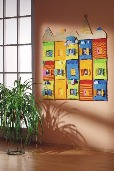 Ein farbenfroher Wandbehang aus Stoff in Form eines Gebäudes mit mehreren Fenstern, von denen jedes ein Kinderfoto zeigt, ist an einer pfirsichfarbenen Wand neben einem großen Fenster und einer grünen Topfpflanze angebracht.