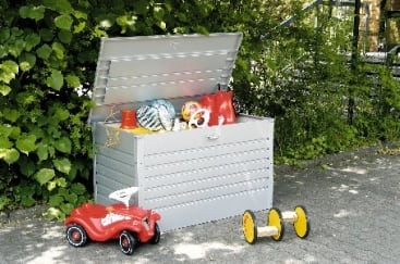 Eine weiße Aufbewahrungsbox für den Außenbereich mit offenem Deckel enthält Spielzeug und Sportgeräte. Vor der Kiste stehen ein rotes Aufsitzauto und eine gelb-schwarze Spielzeugschubkarre, im Hintergrund ist Grün zu sehen.