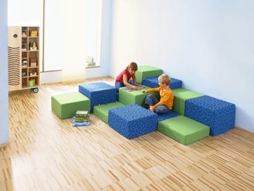 Zwei kleine Kinder sitzen auf grünen und blauen Schaumstoffblöcken, die als Sitzgelegenheiten in einem hellen Raum mit hellem Holzfußboden und blauen Wänden angeordnet sind. Im Hintergrund sind Regale mit Büchern und anderen Gegenständen zu sehen.