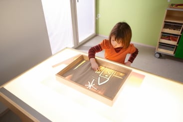 Ein Kind, das ein orangefarbenes Hemd trägt, zeichnet mit dem Finger Formen und Linien in eine Schale mit Sand, die auf einem hellen Tisch in einem hellen Raum steht.