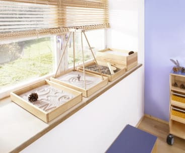 Drei mit Sand und Tannenzapfen gefüllte Holztabletts stehen auf einer Fensterbank unter horizontalen Jalousien. Ein Tablett enthält einen Stock, mit dem man Muster in den Sand zeichnen kann. Das Sonnenlicht fällt durch das Fenster ein.