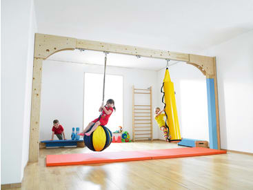 Drei Kinder spielen in einer hellen Turnhalle. Ein Kind schaukelt auf einem großen Ball, ein anderes sitzt auf einer gelben Schaukel in Form eines Bleistifts und das dritte Kind steht auf einem blauen Schwebebalken. Der Boden ist aus Holz und mit roten Matten ausgelegt.