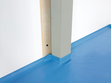 Eine Zimmerecke mit einem blauen Bodenbelag, einer hellgrauen Wand und einem vertikalen Balken, der teilweise von einer blassgrünen Schutzhülle bedeckt ist, mit einer Holzplatte, die mit einer sichtbaren schwarzen Schraube befestigt ist.