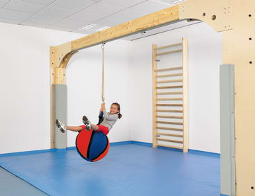 Ein kleines Kind sitzt auf einem rot und blau gefederten Schaukelball in einem Turnraum mit gepolstertem blauem Boden und einer an der Wand befestigten hölzernen Kletterleiter. Das Kind lächelt und hält sich an dem Seil fest.