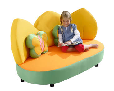 Ein junges Mädchen sitzt im Schneidersitz auf einem bunten, kindgerechten Sofa mit gelben blattförmigen Rückenlehnen und schmetterlingsförmigen Kissen und liest ein Buch. Das Sofa hat einen grünen Sockel und eine orangefarbene Sitzfläche.