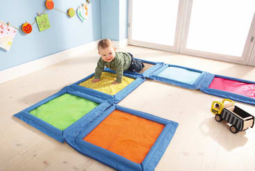 Ein Baby krabbelt auf bunten, strukturierten Matten, die auf einem hellen Holzboden ausgelegt sind. In der Nähe steht ein Spielzeugauto, und im Hintergrund ist eine blaue Wand mit Kinderkunstwerken zu sehen. Helles natürliches Licht fällt durch große Fenster ein.