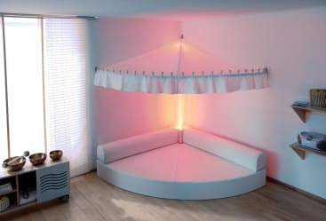 In einem modernen Raum mit hellem Holzboden steht ein Ecksofa mit weißen Kissen und einem großen weißen Baldachin über dem Kopf. Sanftes rosa Licht erhellt die Ecke. In der Nähe befinden sich Regale und ein Schrank mit Schüsseln und einer Stereoanlage.