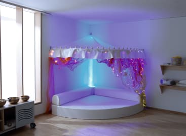 Ein weißer, halbkreisförmiger, gepolsterter Sitzbereich befindet sich in der Ecke eines sanft beleuchteten Raums, der teilweise von einem Stoffbaldachin mit violetten und goldenen Lichterketten umgeben ist. Im Hintergrund sind Regale und große Fenster zu sehen.