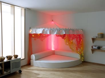 Ein weißes, halbrundes, gepolstertes Sofa steht in einer Ecke unter einem Baldachin aus orangefarbenem und rosafarbenem Stoff und Lichterketten, die von rosafarbenen Lampen beleuchtet werden. In der Nähe sind Regale und Holzschalen zu sehen.