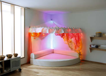 Ein weißer, halbkreisförmiger, gepolsterter Sitzbereich mit einem mit orangefarbenem Stoff bespannten Baldachin und Lichterketten, an einer weißen Wand in einem Raum mit Holzboden und Regalen mit verschiedenen Gegenständen.