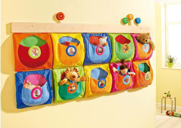 An der Wand montierter Organizer mit zehn bunten Stofftaschen, die jeweils ein kleines Stofftier oder Spielzeug enthalten. Die Taschen sind in zwei Fünferreihen angeordnet. Die Wand ist hellgelb, und auf einer Fensterbank ist eine Pflanze zu sehen.