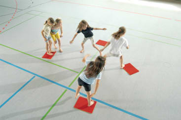 Fünf Kinder stehen barfuß auf roten Matten, die auf einem mit farbigen Linien markierten Turnhallenboden liegen, und scheinen ein Gruppenspiel zu spielen. Die Szene ist hell beleuchtet, und alle Kinder schauen in verschiedene Richtungen.