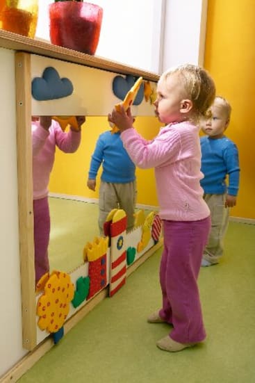 Ein kleines Kind in einem rosafarbenen Outfit steht vor einem Spiegel und hält ein Spielzeug, während ein anderes Kind in einem blauen Hemd und einer beigen Hose im Hintergrund steht. Der Spiegel ist mit bunten Formen verziert.