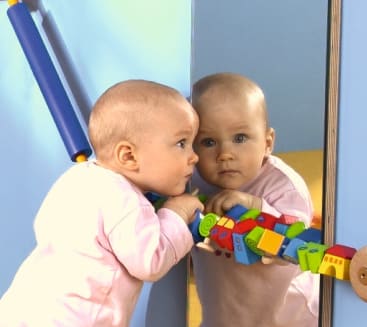 Ein Baby in einem rosa Outfit lehnt an einem Spiegel und hält ein buntes Holzspielzeug mit verschiedenen Formen. Im Spiegel ist das Spiegelbild des Kindes zu sehen. Der Hintergrund ist blau und gelb.