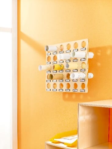 An einer gelben Wand neben einem Fenster ist ein hölzerner Wandorganizer mit mehreren runden Schlitzen angebracht, die jeweils beschriftet sind und aufgerollte Papiere oder Poster aufnehmen. Unter dem Organizer befindet sich ein Regal mit Ordnern.