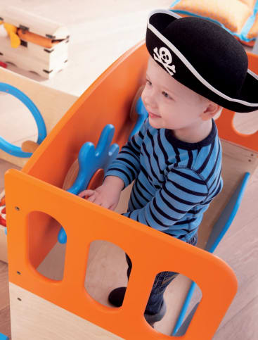 Ein kleines Kind, das einen schwarzen Piratenhut mit einem Totenkopf trägt, steht am Lenkrad eines bunten Spielgeräts, schaut nach vorne und lächelt. Das Kind ist mit einem blau-schwarz gestreiften Hemd bekleidet.