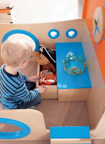 Ein kleines Kind sitzt in einer hölzernen Spielstruktur und greift in eine Kiste mit Plüschtieren. Ein Piratenhut und eine grüne Krone sind in der Nähe auf blauen Flächen platziert. Die Umgebung ist mit orangefarbenen und blauen Akzenten versehen.