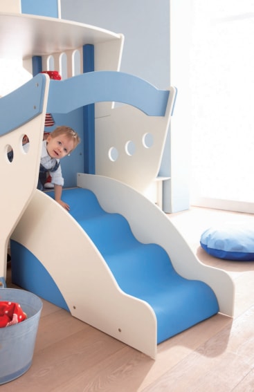 Ein kleines Kind steht oben auf einer blau-weißen Indoor-Rutsche, die an einem Spielgerät in einem hell erleuchteten Raum mit Holzboden befestigt ist. In der Nähe befinden sich ein blaues Kissen und ein Metalleimer mit Spielzeug.