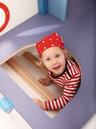Ein kleines Kind, das ein rot-weiß gepunktetes Kopftuch und ein rot gestreiftes Hemd trägt, schaut nach oben, während es durch eine kreisförmige Öffnung in einem Spielgerät mit lila und weißen Oberflächen klettert.