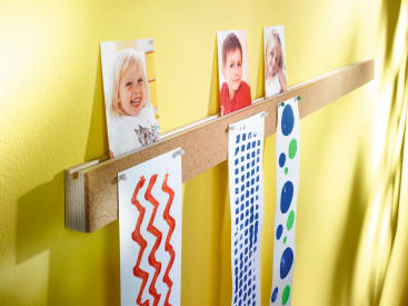 In einem hölzernen Wandregal sind drei Kinderfotos und ihre farbenfrohen Kunstwerke, darunter rote Schnörkel und blaue Punktmuster, vor einer gelben Wand ausgestellt.