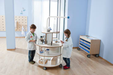 Zwei junge Kinder in weißen Laborkitteln stehen sich in einem hell erleuchteten Klassenzimmer mit hellem Holzfußboden und blauen Wänden gegenüber, in dem ein runder, rollender Wissenschaftswagen mit verschiedenen Laborgeräten steht.