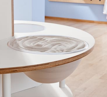 Ein runder weißer Tisch mit einer eingebauten durchsichtigen Schale, die mit glattem, aufgewirbeltem Sand gefüllt ist, steht auf einem hellen Holzboden neben einer blauen Wand.